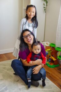 Educator sitting on the floor holding a baby while an older child stands behind them in a bright playroom, representing a nurturing daycare for 1 year old children in a safe, supportive early learning environment.